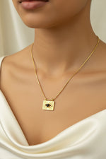 Load image into Gallery viewer, A gold-plated stainless steel necklace with a geometric eye-shaped pendant and snake type chain detailing, displayed on a mannequin against a green backdrop with white silk flowers.
